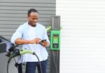 An African man is standing near an electric car, waiting for it to charge at a charging station and using the phone