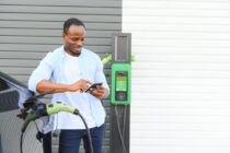 An African man is standing near an electric car, waiting for it to charge at a charging station and using the phone