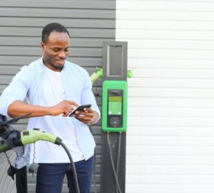 An African man is standing near an electric car, waiting for it to charge at a charging station and using the phone
