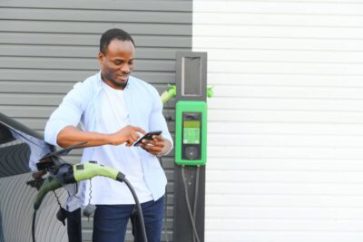 An African man is standing near an electric car, waiting for it to charge at a charging station and using the phone
