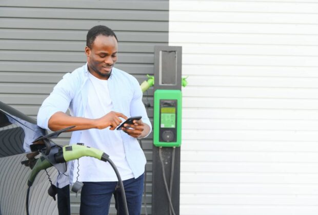 An African man is standing near an electric car, waiting for it to charge at a charging station and using the phone