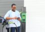An African man is standing near an electric car, waiting for it to charge at a charging station and using the phone